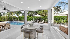 A modern indoor pool area with a round table and chairs, a seating area with lounge chairs and umbrellas, and a view of a tropical garden. - Gorgeous Modern Beach Heated Pool House Sleeps 8 Central (Delray Beach)