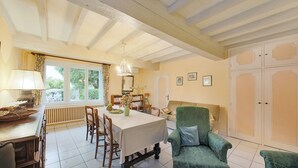 Ein Essbereich mit Tisch, Stühlen und einem grünen Sessel. Ein Fenster mit Vorhängen und ein Heizkörper unter dem Tisch sind ebenfalls zu sehen.
