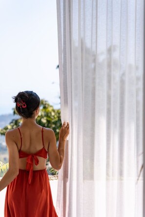 A person in a red dress standing by a window with sheer curtains, overlooking a view of greenery and a body of water.