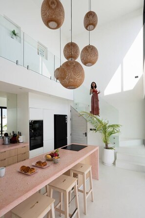A modern kitchen with a pink countertop, wooden stools, and hanging wicker light fixtures.