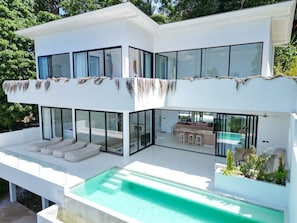Una casa moderna de dos pisos con piscina, ventanas de vidrio amplias y mobiliario de exterior.