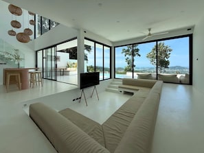 A modern living room with a large sectional sofa, a flat-screen TV on a stand, a dining area with a table and chairs, and large windows offering a view of the outdoors.