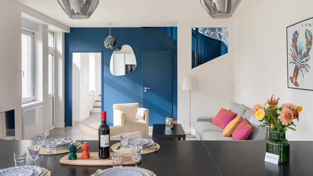 Een moderne woonkamer met een blauwe accentmuur, een eethoek met een tafel gedekt voor twee personen en een trap naar een hoger niveau.