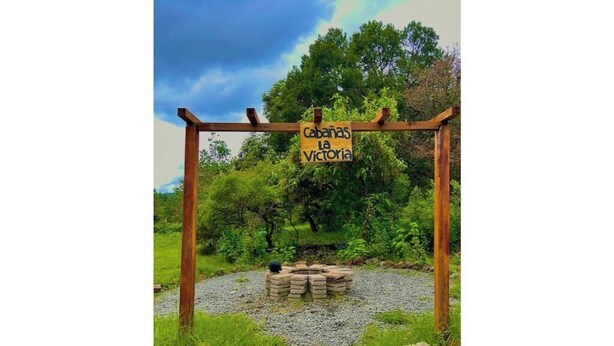Una pérgola de madera con un letrero que dice "Cabanas La Victoria" en un entorno natural con árboles y césped.