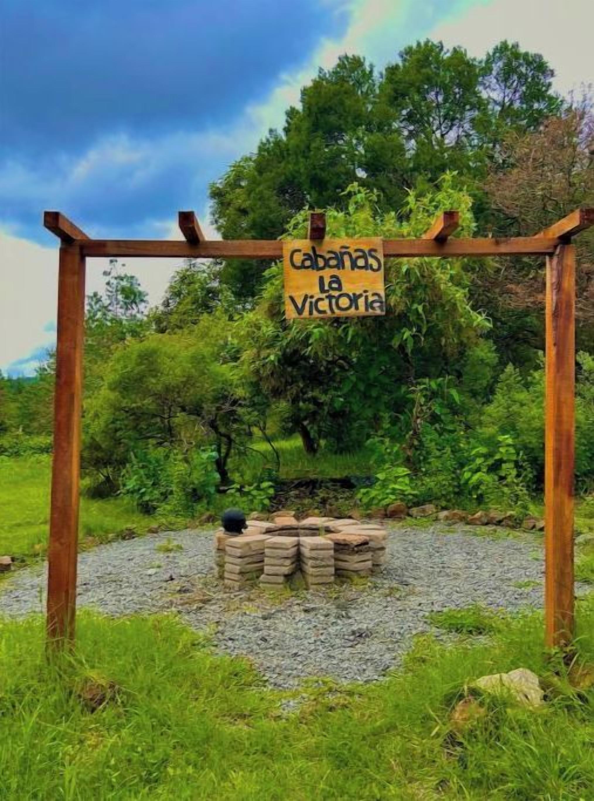 A wooden pergola with a sign reading "Cabanas La Victoria" in a natural setting with trees and grass.