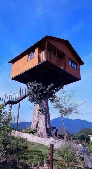 A treehouse with a wooden deck and a staircase leading up to it, surrounded by greenery and a mountain in the background.