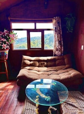 A cozy room with a large window, a cushioned sofa, a glass coffee table, and a potted plant. - Trini Tree House — Nido By GEstores (Omitlán de Juárez)
