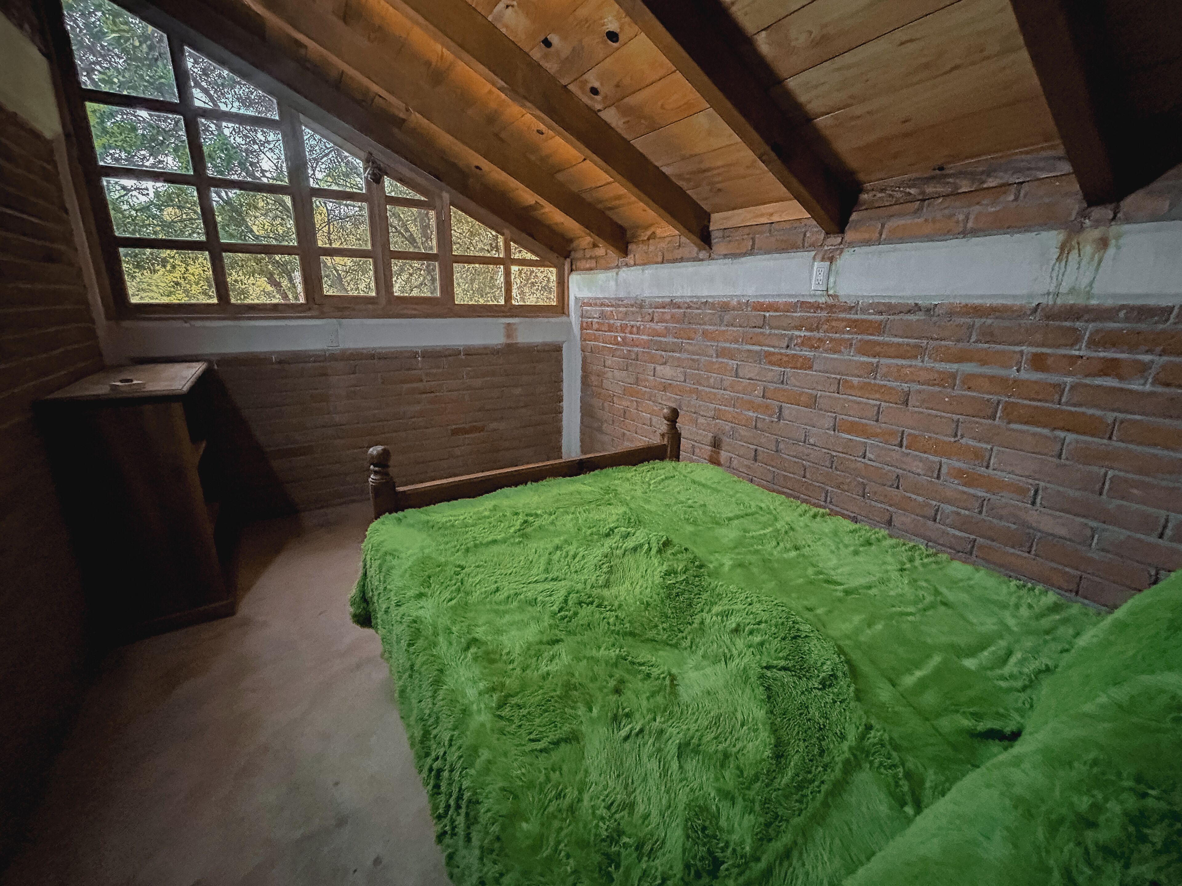 Een kamer met een houten plafond, een bakstenen muur, een raam, een bed met een groene deken en een kleine tafel.