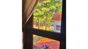 A view through a window with a wooden frame, revealing a red-roofed building and a tree-lined yard.