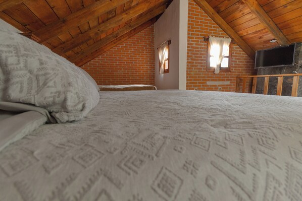 Una habitación con techo de madera inclinado, pared de ladrillo, cama con ropa de cama estampada, ventana pequeña con cortinas y televisor montado en la pared.