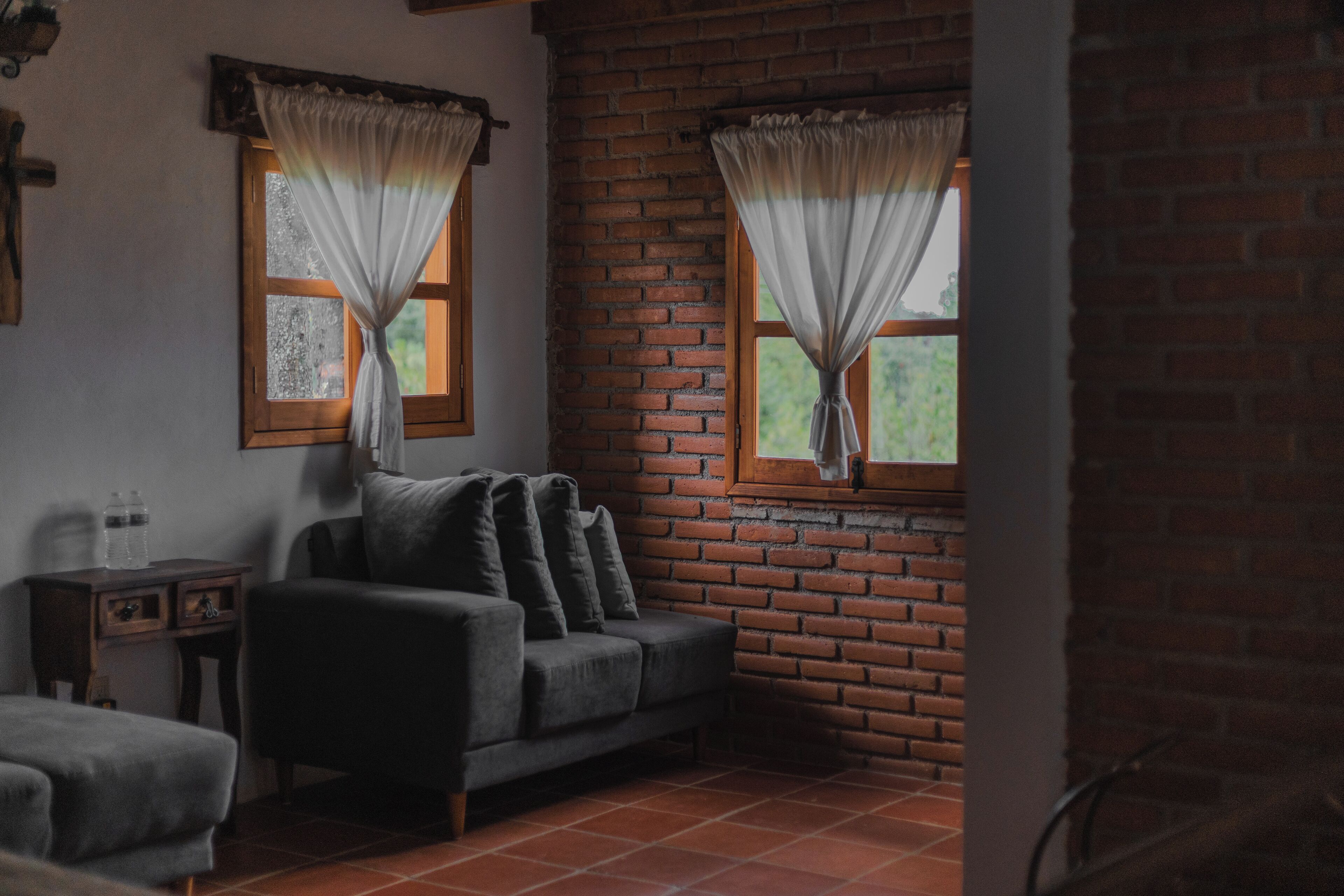 Una habitación acogedora con pared de ladrillo, dos ventanas con cortinas, un sofá y una mesita con una botella de agua.