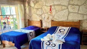 Dos camas individuales con ropa de cama azul y almohadones decorativos, una pared de piedra y una ventana con vistas a la vegetación.