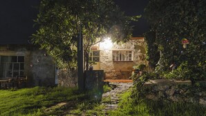 Un bâtiment en pierre avec une fenêtre illuminée, entouré de verdure et bordé d’un chemin en pierre.