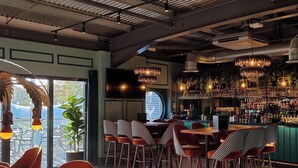 A bar area with a high ceiling, exposed ductwork, and a variety of hanging light fixtures. There are multiple seating areas with checkered tablecloths and bar stools.