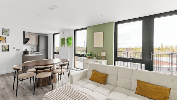 A modern living room with a sofa, a dining table, and chairs. There is a kitchen area in the background with a window overlooking a balcony.