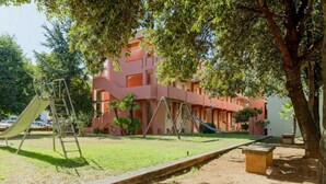 A playground with slides and swings, a building with a pink facade, and trees providing shade.