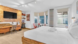 Une chambre d’hôtel moderne dotée d’un grand lit, d’un bureau avec un ordinateur, d’un téléviseur à écran plat fixé à un mur en briques et de grandes fenêtres avec des rideaux.