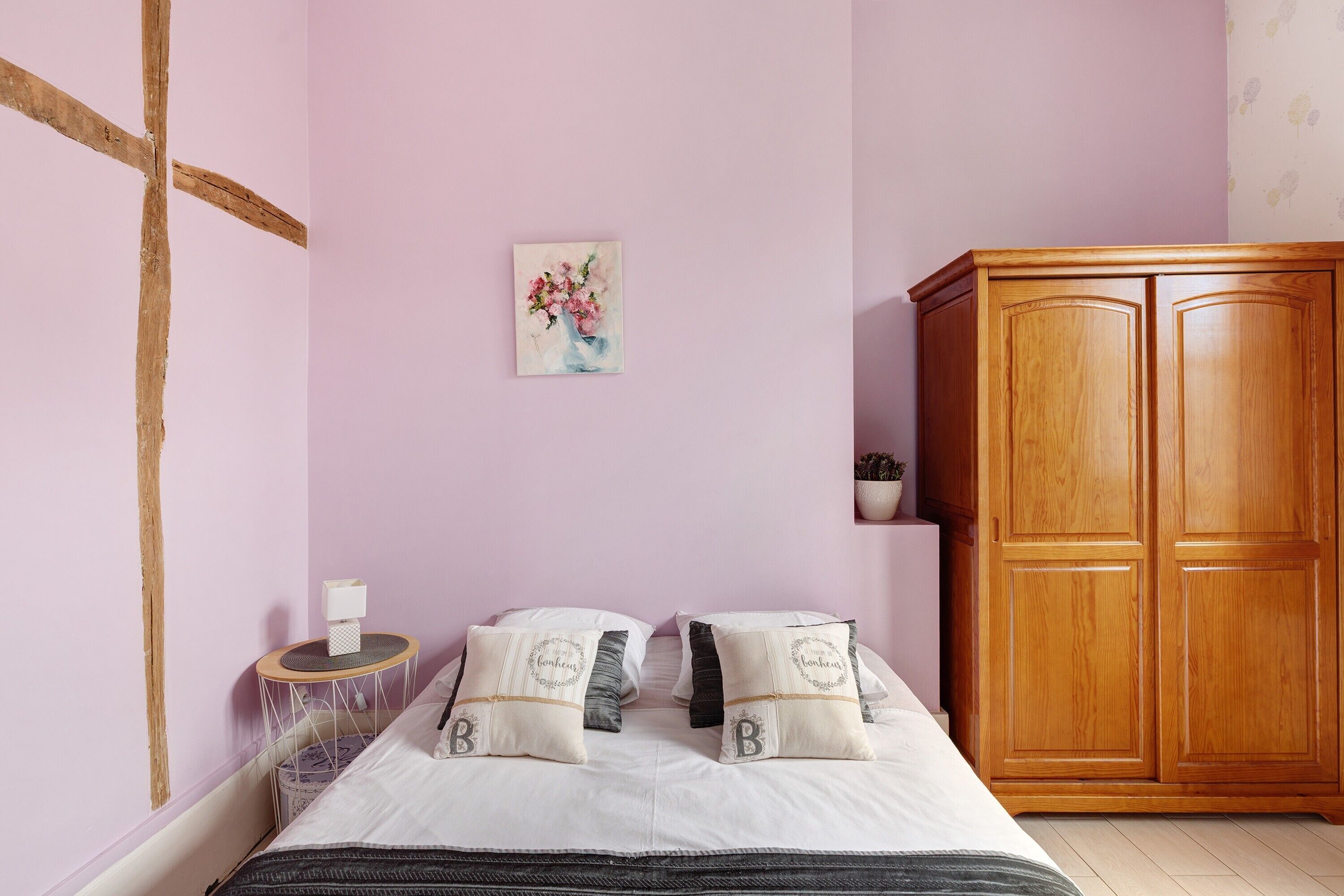 A bedroom with a bed, pillows, a wooden wardrobe, a small round table, and a wall-mounted painting.