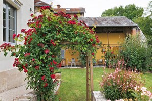 Un jardí amb una pèrgola de fusta coberta de flors vermelles, un edifici groc i un prat verd.