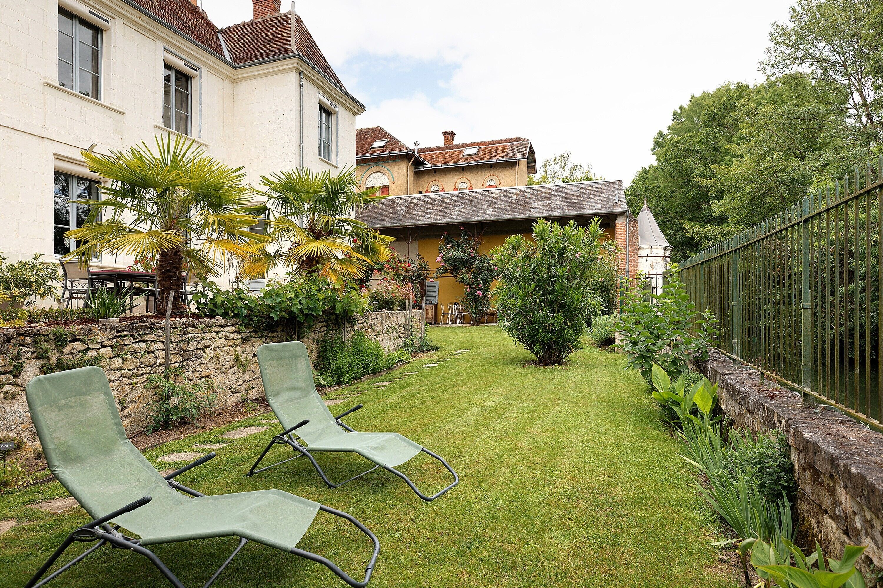 A well-maintained garden with two lounge chairs, a stone wall, and a building with a tiled roof.