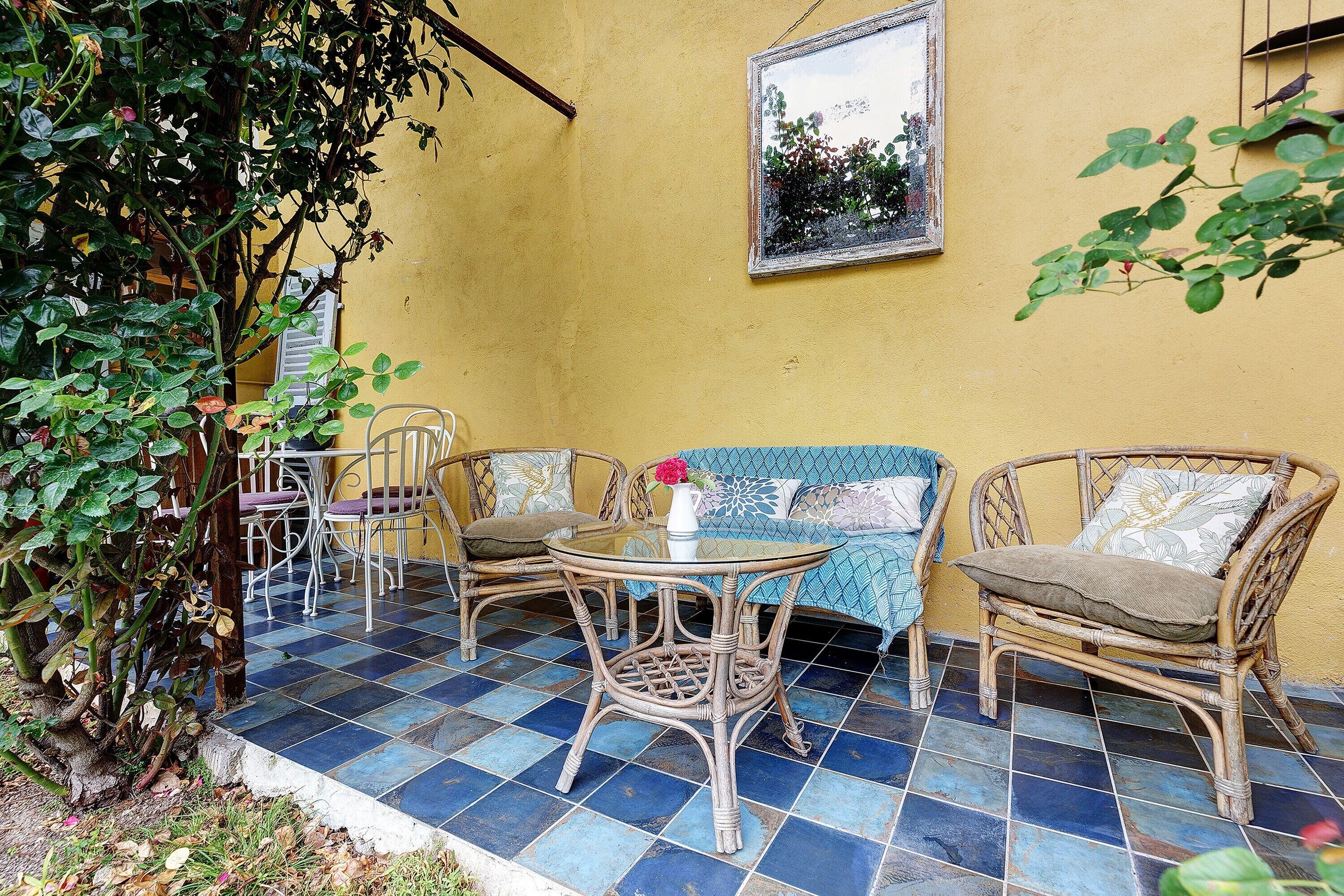 Un patio con mobiliario de mimbre, una mesa de vidrio y una pared amarilla.