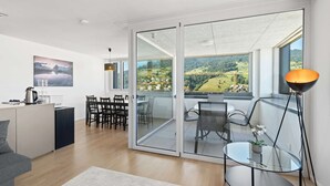 A modern living room with a dining area, a kitchen, and large glass doors leading to a balcony with a view of a valley.