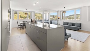A modern kitchen with a central island, dining area, and a sofa in a bright room with large windows.