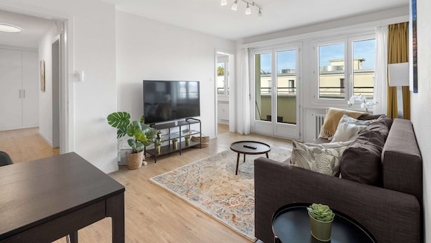 Ein modernes Wohnzimmer mit Flachbildfernseher, einer Couch, einem Couchtisch und einem Esstisch. Ein großes Fenster bietet Blick auf Gebäude und es gibt einen Balkon.
