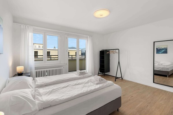 Un dormitorio moderno con una cama grande, una mesita de noche con lámpara, una ventana con cortinas, un armario negro y un espejo grande en la pared.