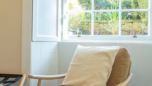 Een kamer met een raam dat uitkijkt op een tuin, een houten stoel met een beige kussen en een klein tafeltje met een blauw-witte streep tafelkleed.
