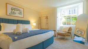 Een netjes opgemaakt bed met een blauw hoofdeinde, een stoel, een ladder en een raam met uitzicht op groen.
