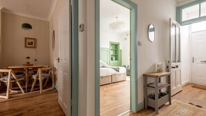 A room with a wooden floor, a dining table with chairs, a small table with a lamp, and a bed with a green cabinet.