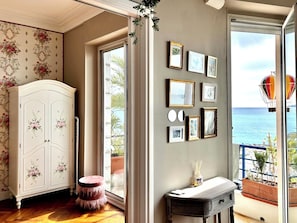 A room with a white cabinet with floral designs, a round stool, a small table with a bottle, and a wall with framed pictures. - PANORAMIC SEA VIEW | Luxury Design for 4 | DOWNTON ABBEY Style (Nice)