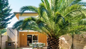 A modern house with a wooden exterior, a patio with a table and chairs, and a large palm tree. - Holiday Home 'Maison De Vacances Oceanides' with Private Terrace, Wi-Fi and Air Conditioning (Capbreton)