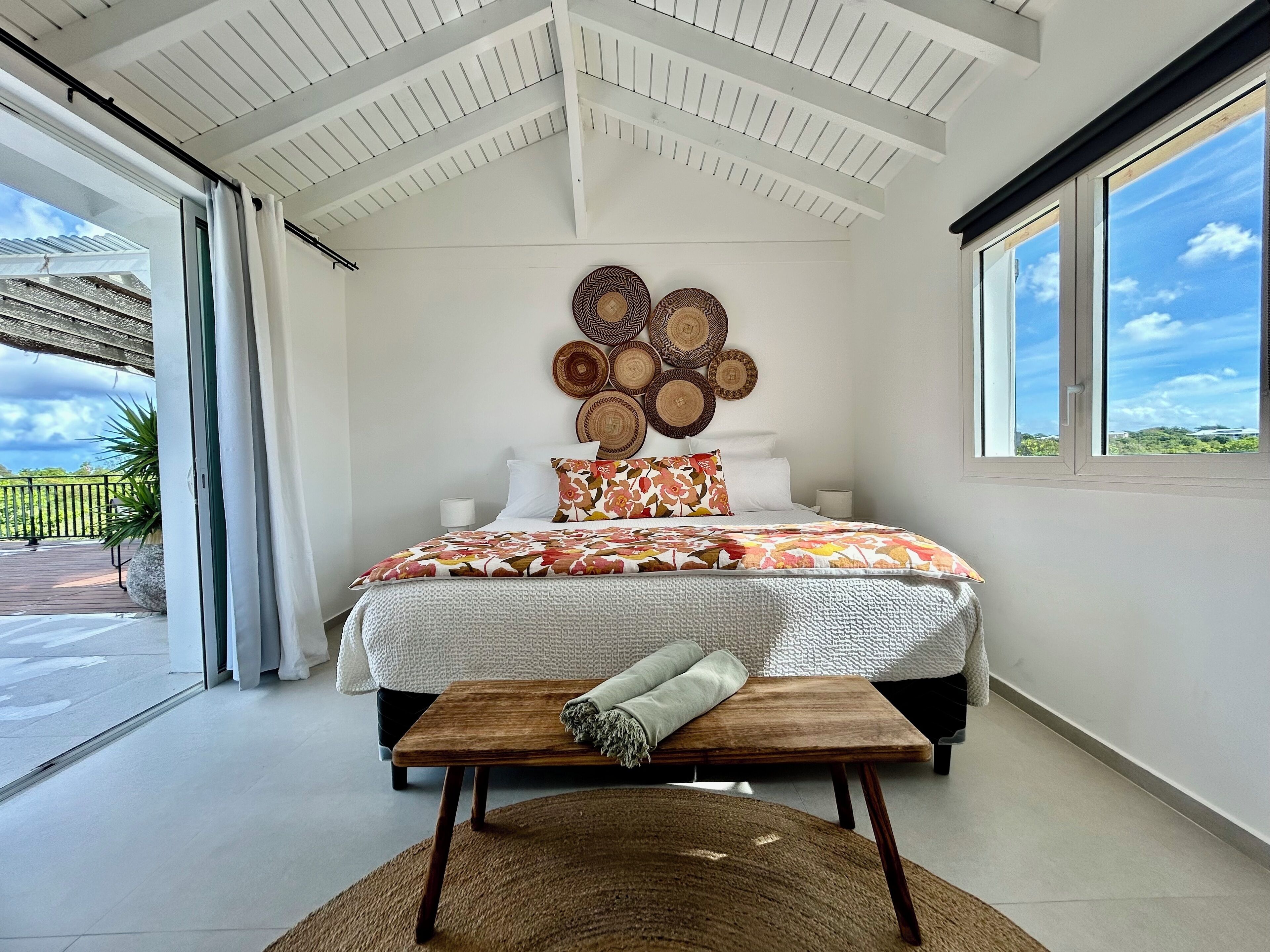 Ein Schlafzimmer mit einem großen Bett, einer hölzernen Bank, einem gewebten Teppich und Blick ins Freie durch ein Fenster.