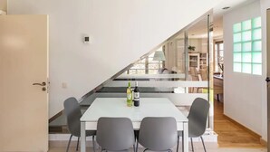 Un espace repas moderne avec une table blanche et quatre chaises, une bouteille de vin et un escalier menant à un niveau supérieur.