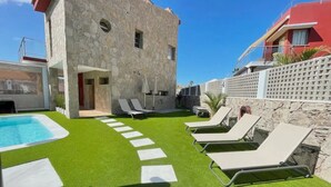 Un espace piscine agrémenté de gazon synthétique, de chaises longues et d’un bâtiment en pierre.