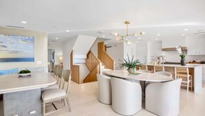 A modern kitchen with white cabinets, a central island, and a dining area with round tables and chairs. - Siesta Beachfront Paradise - Infinity Pool & Spa! (Sarasota)