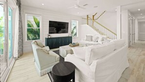 A spacious living room with a white sofa, a coffee table, and a TV mounted on the wall. A staircase with a wooden railing is visible in the background. - Beach View Paradise - New Anna Maria Island Home (Bradenton Beach)