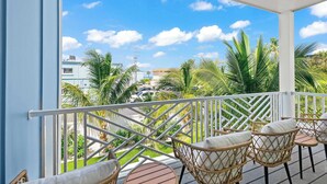 A balcony with wicker chairs and a table, overlooking a street with palm trees and buildings. - Beach View Paradise - New Anna Maria Island Home (Bradenton Beach)