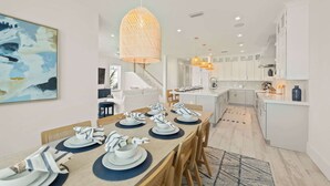 A modern kitchen with white cabinets, a central island, and a dining area set for six with striped napkins and plates. - Beach View Paradise - New Anna Maria Island Home (Bradenton Beach)