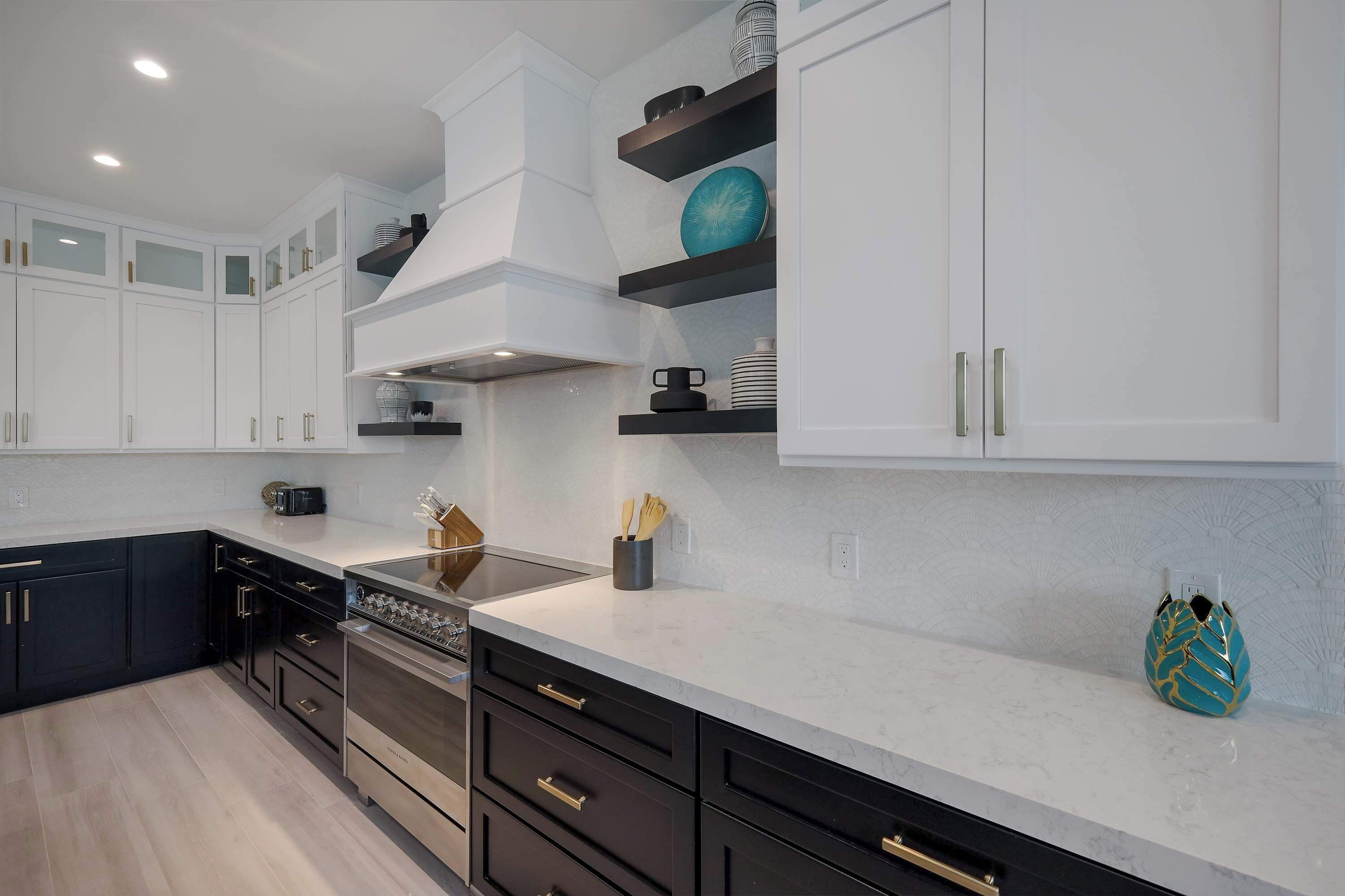 A modern kitchen with white cabinets, black lower cabinets, and a white countertop. There are built-in shelves with decorative items and a stove.