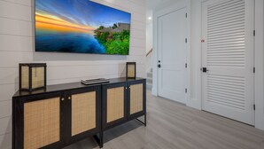 A modern hotel room with a large flat-screen TV mounted on the wall, a dark wooden console table with rattan panels, and a white door with horizontal louvered panels.