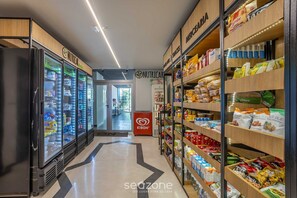 Interieur van een supermarkt met koelvitrines, planken vol snacks en levensmiddelen, en een Kibon logo op een toonbank.