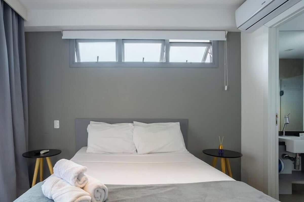 Une chambre dâhĂŽtel moderne, Ă©quipĂ©e dâun lit, de deux tables de chevet, dâune fenĂȘtre et dâune salle de bain visible Ă travers une porte ouverte.