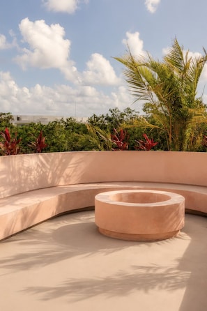 Un espacio de descanso al aire libre con forma curva, que cuenta con una jardinera circular en el centro, rodeada de follaje y palmeras.
