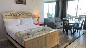A hotel room with a bed, a small table, a sofa, and a balcony with chairs. - Cozy Ocean Front Getaway ~ Sleeps 2 ~ Pools Open! (Daytona Beach)
