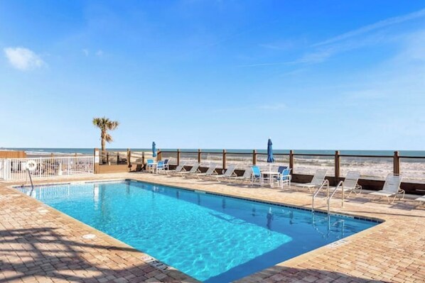 A pool with clear blue water, surrounded by lounge chairs and umbrellas, with a beach and ocean in the background.