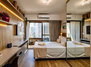 A modern hotel room with a large bed, a desk, a mirror, and a view of the city.