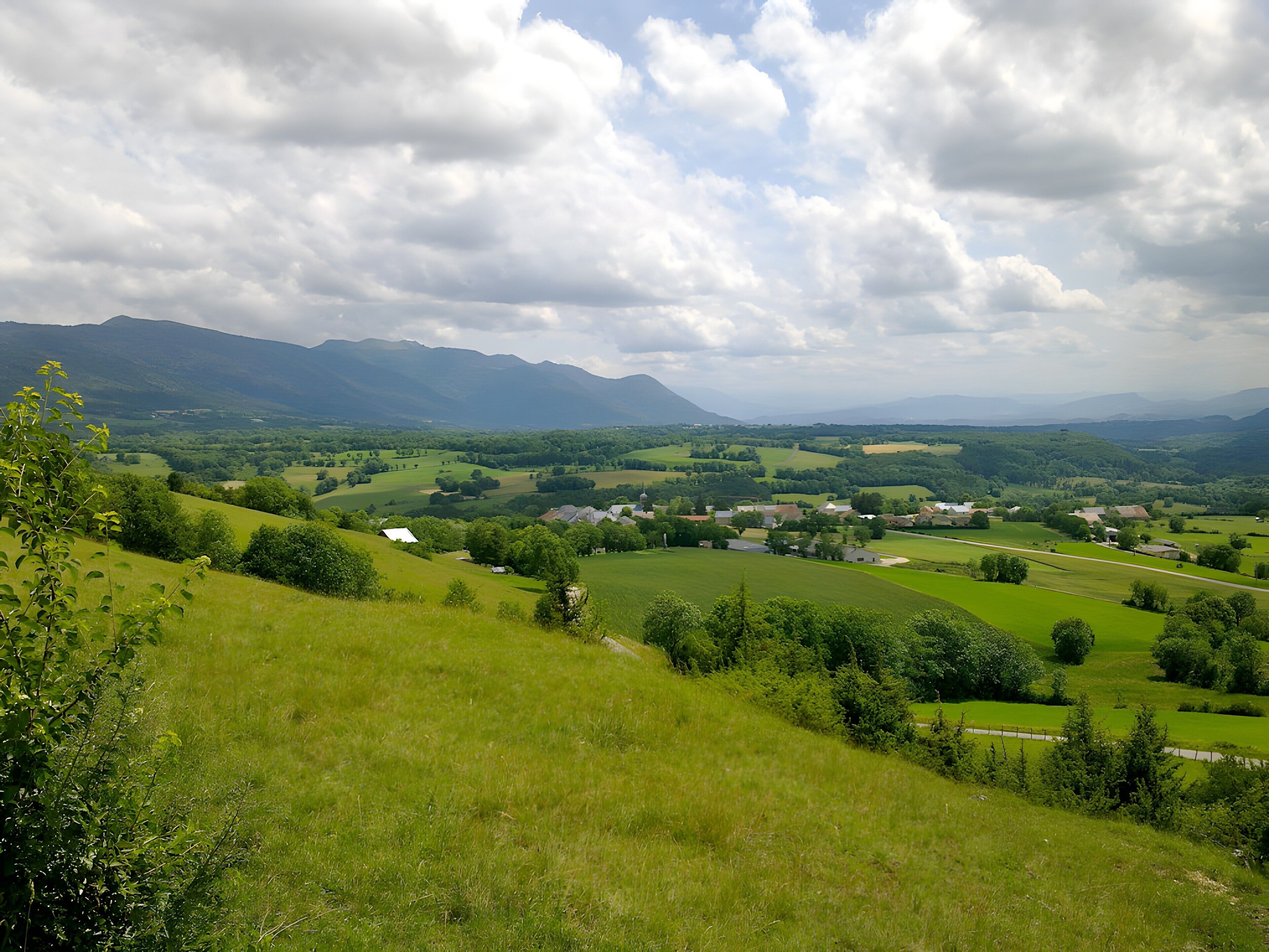 Un paysage rural caractérisé par des collines douces, des maisons éparpillées et des montagnes à l’horizon.